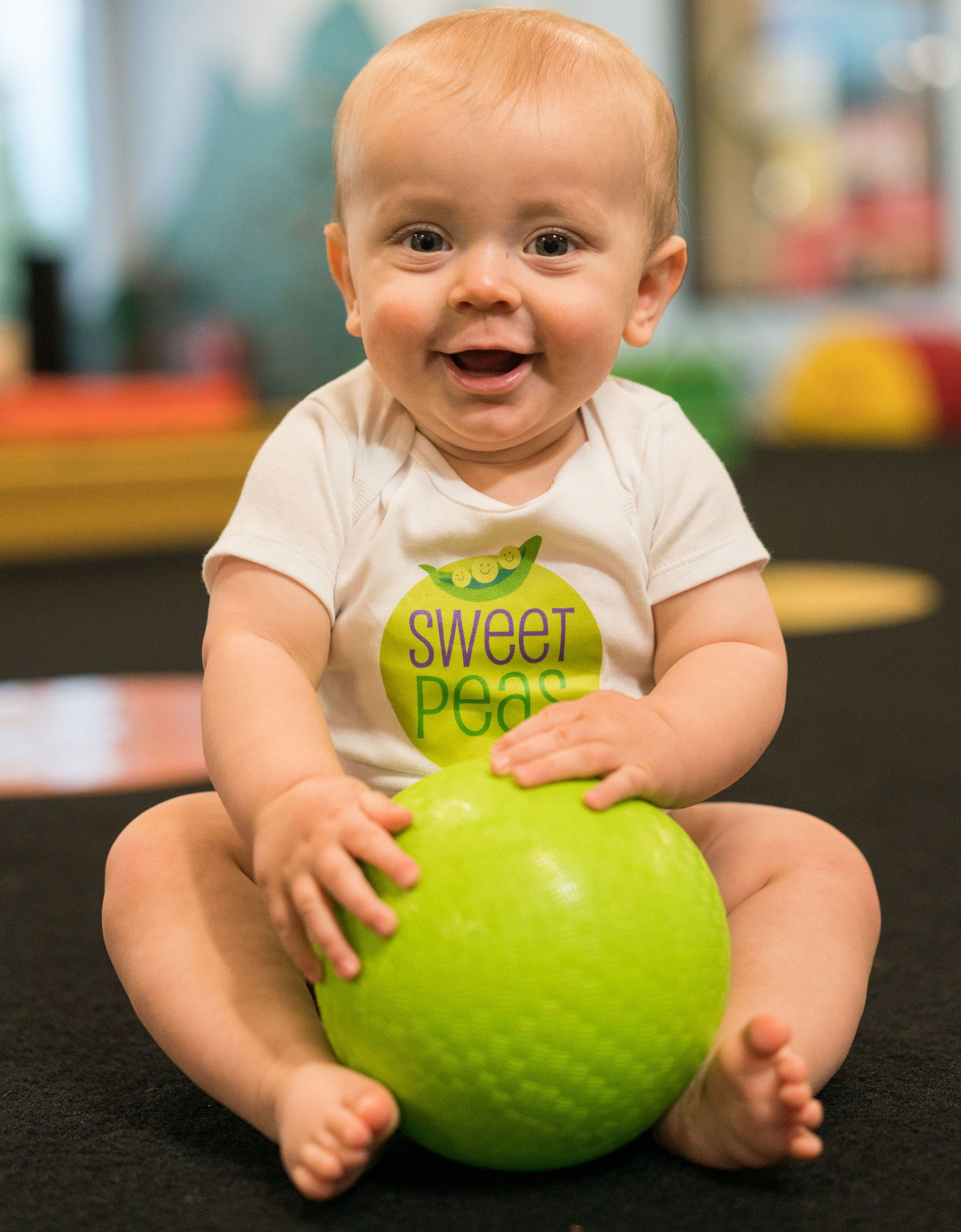 Blonde child smiles, crawling on a green surface toward the viewer in a pit filled with foam cubes.