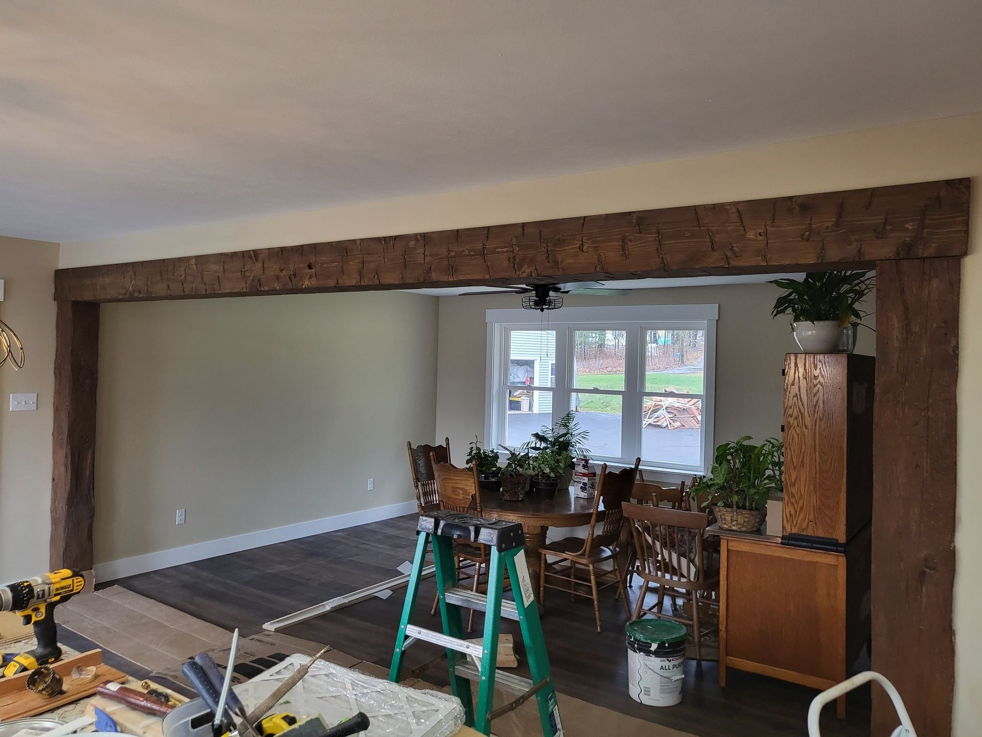 Living room renovation with exposed ceiling beam, ladders, tools, and plants near a window