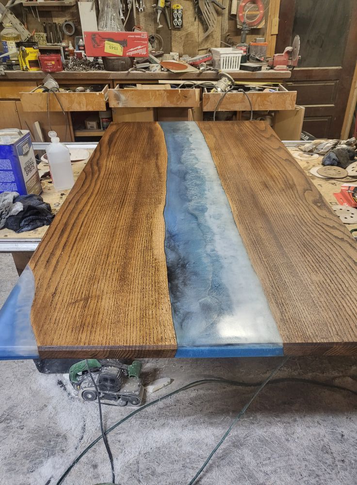 Wooden table slab with blue resin center in a cluttered workshop, clamped on a workbench.