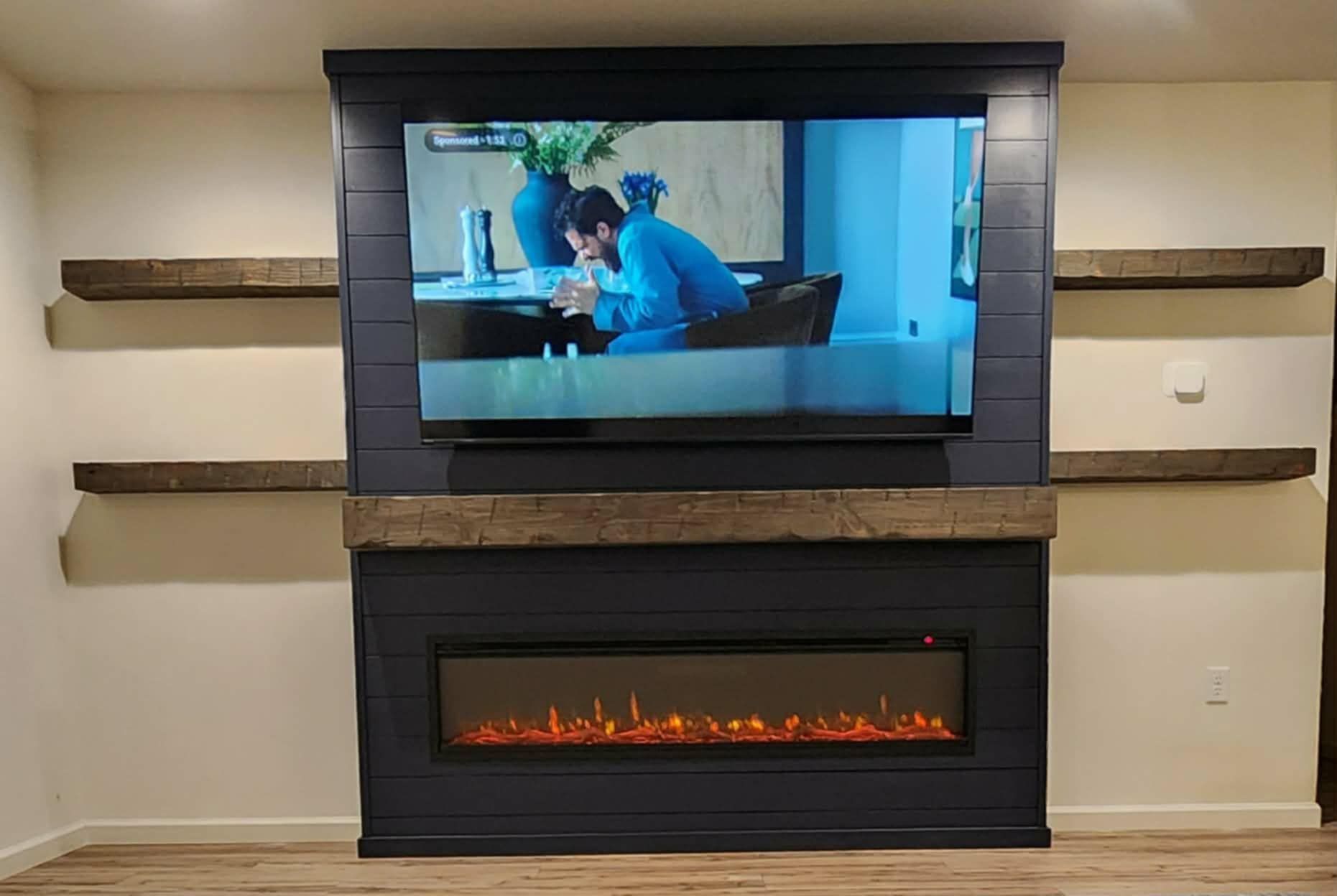 Wall-mounted TV above a modern electric fireplace with wood mantel and floating shelves on a beige wall.