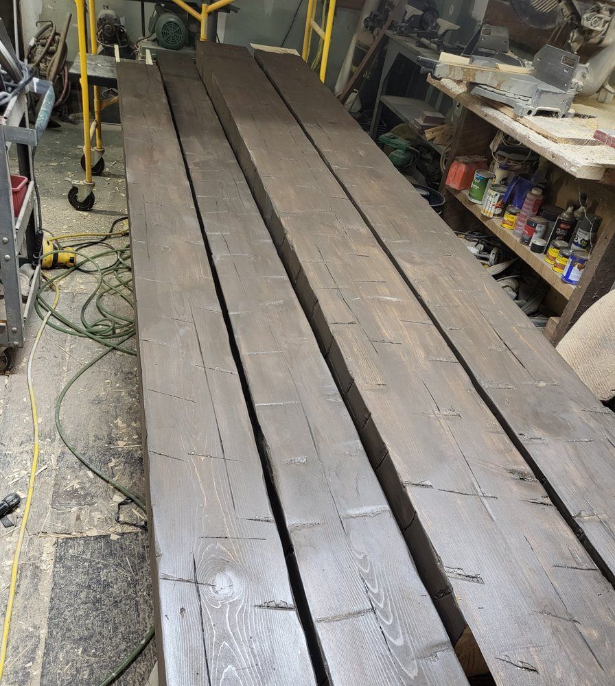 Long wooden planks laid out in a cluttered workshop aisle, with tools and shelves nearby.