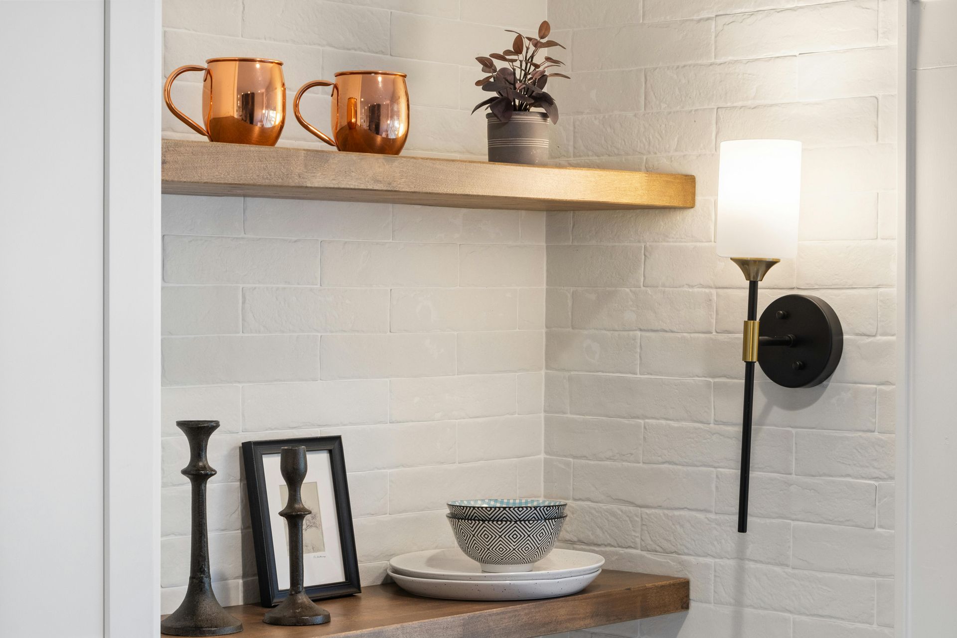 White shelf and wall sconce above a wooden console table with mugs, candles, and decor in a white brick nook