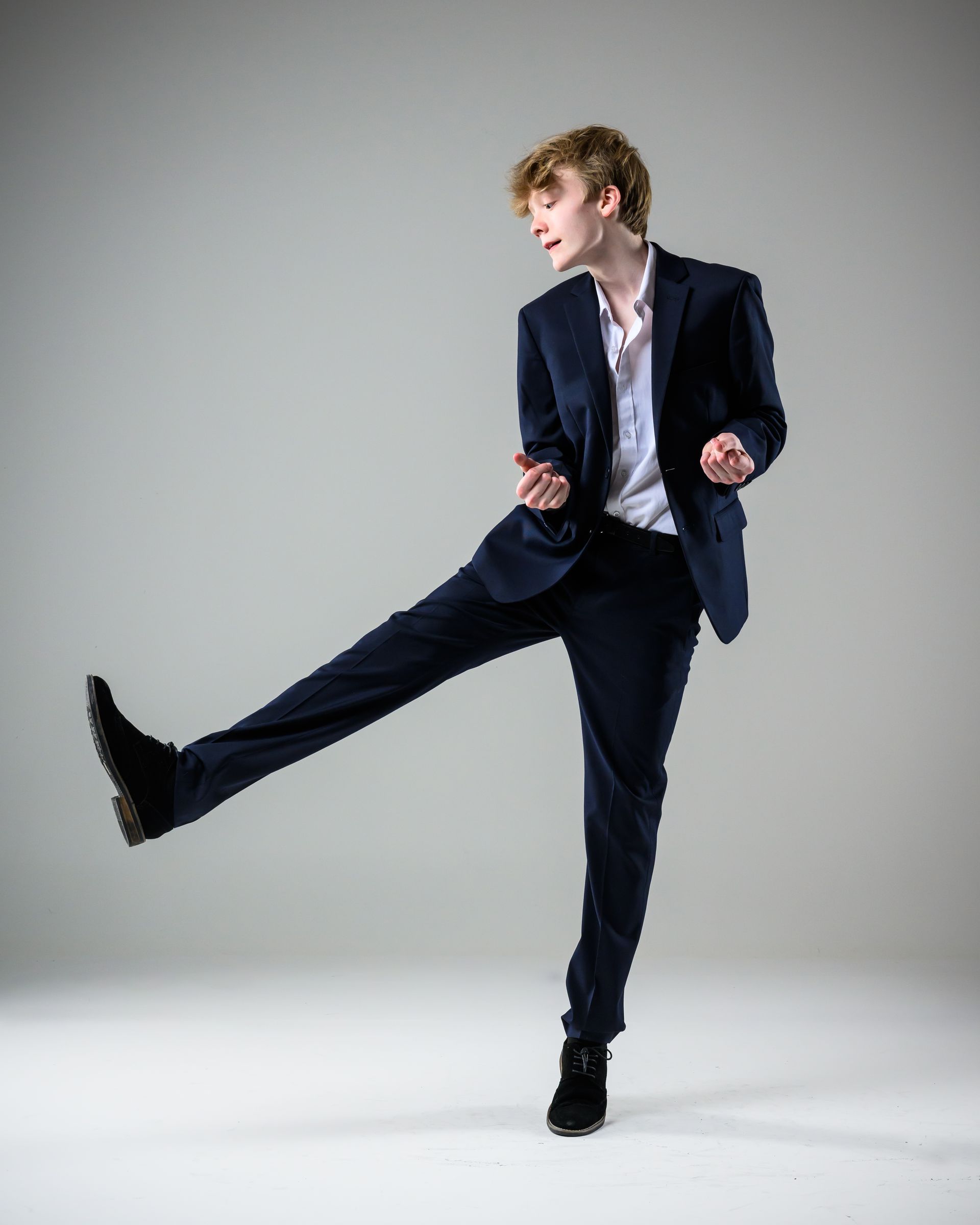 Person in a dark suit kicks a leg out against a gray studio background.
