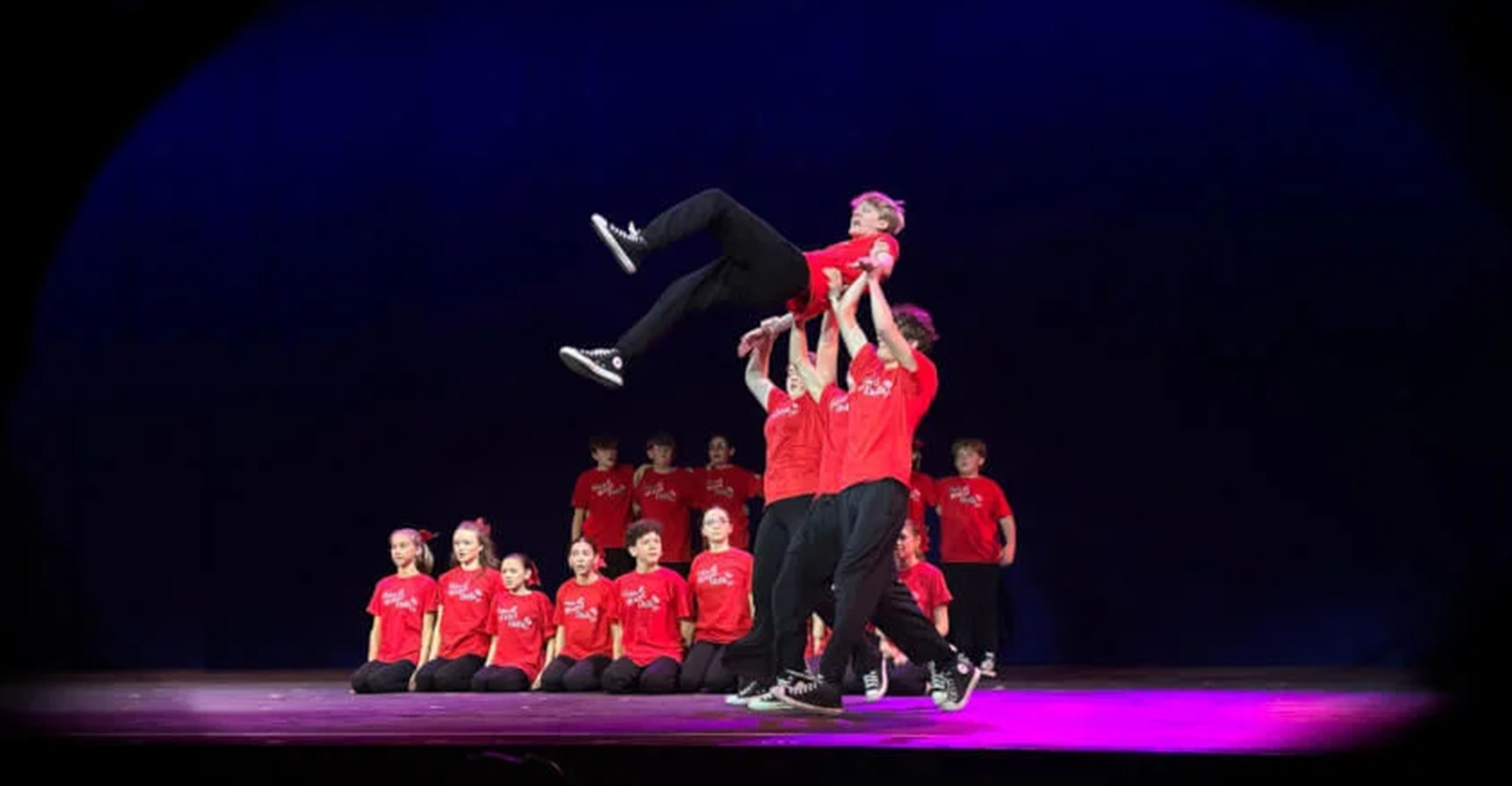 Dancers in red outfits perform a choreographed stage lift under purple lighting