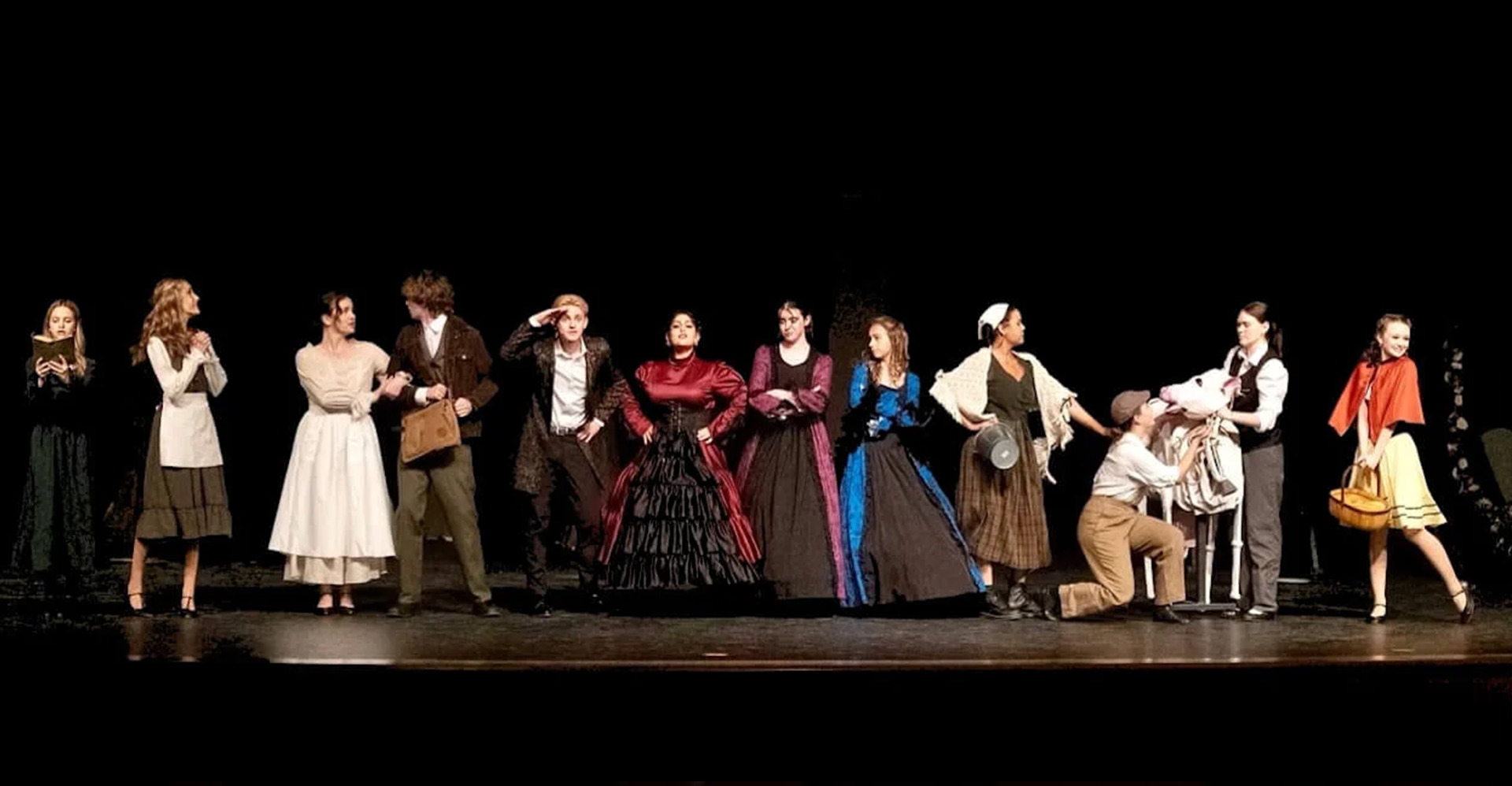 Costumed performers standing in a line on a dark stage, some posing and dancing in bright theatrical outfits