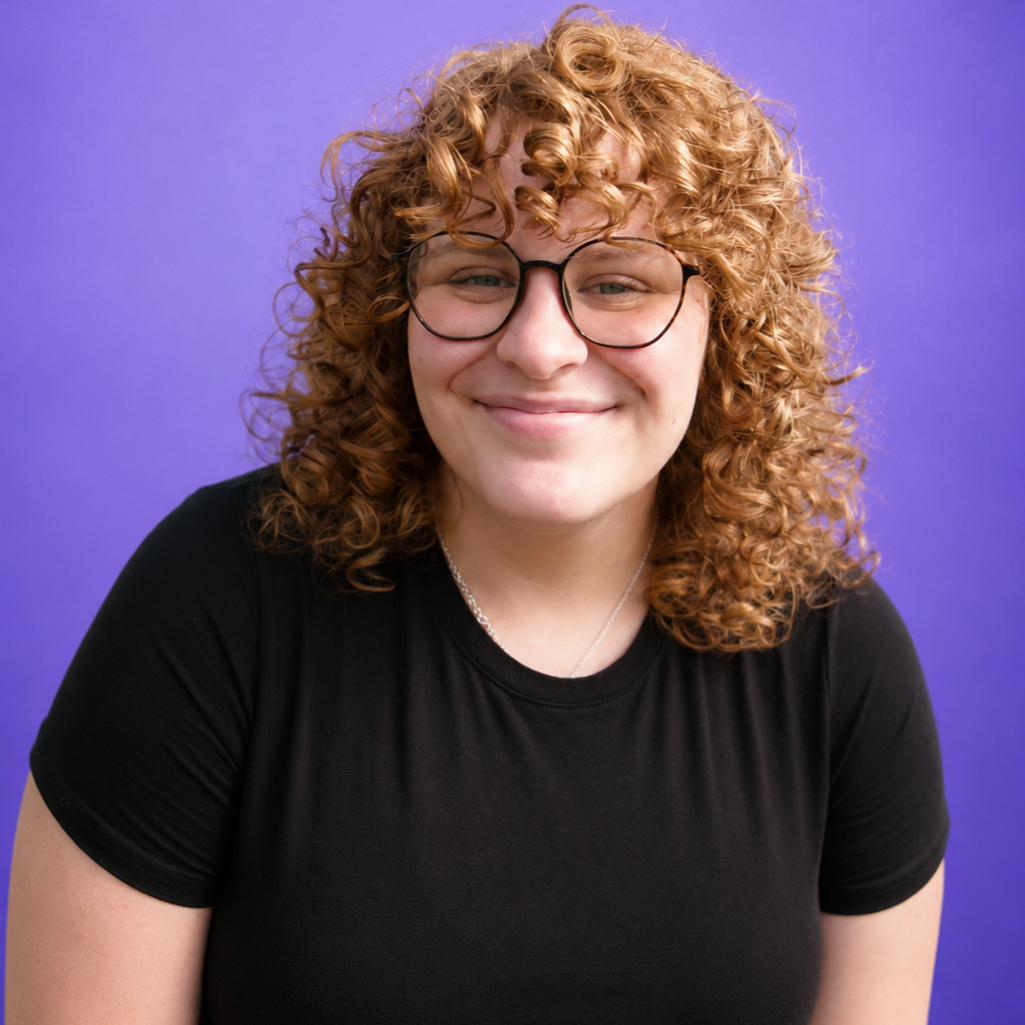 Smiling person with curly hair and glasses wearing a black shirt against a purple background