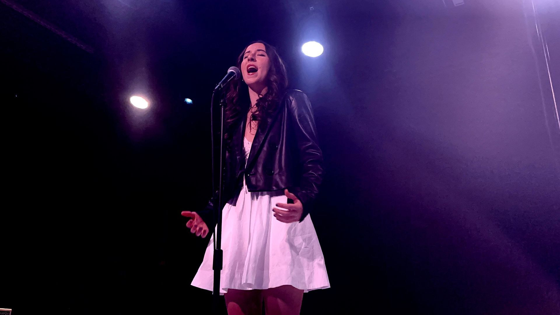 Singer performing onstage under purple spotlights, wearing a black jacket and white skirt, singing into a microphone