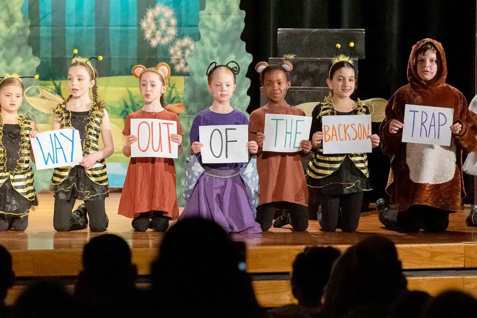 Children in costumes onstage holding signs that spell “WILL OUT OF THE BOX TRAP”