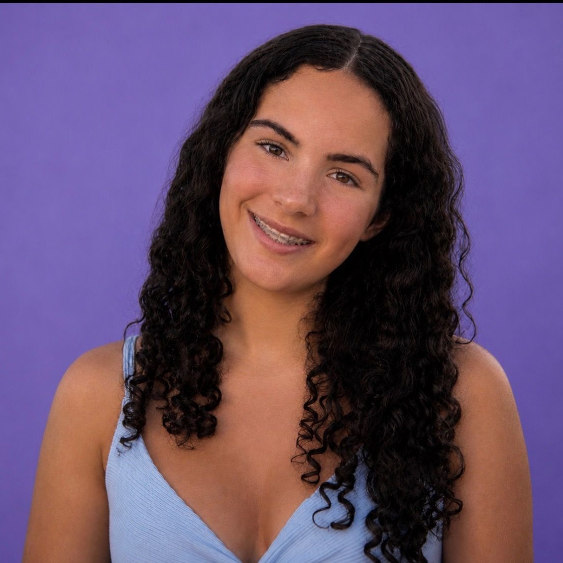 Smiling woman with long curly dark hair against a purple background, wearing a light blue top