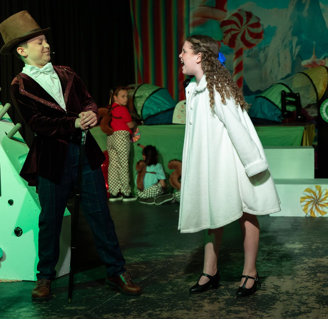 Two performers in colorful costumes face each other on a stage with whimsical set pieces.