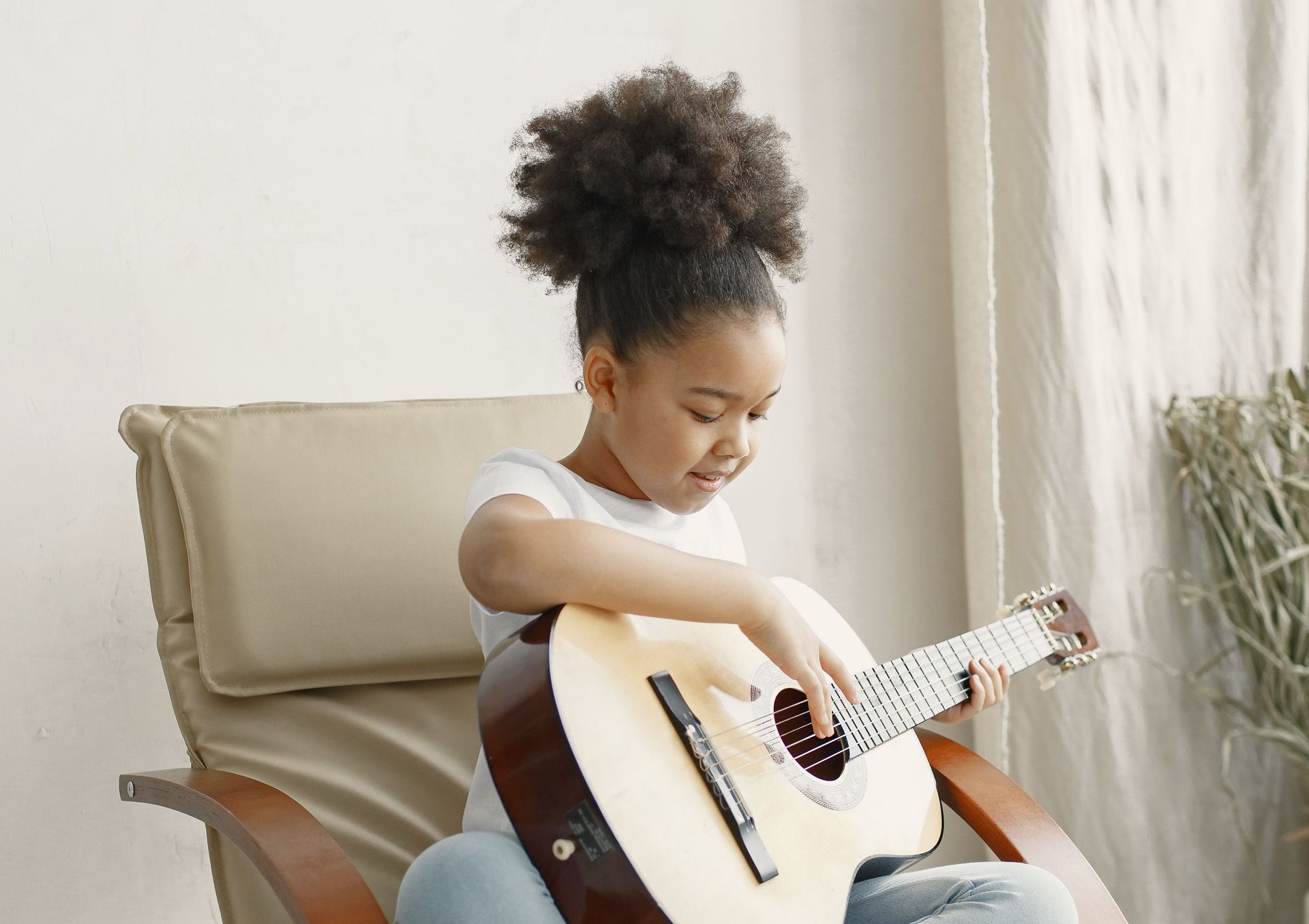 Young child sitting in a chair, playing a light-colored acoustic guitar indoors