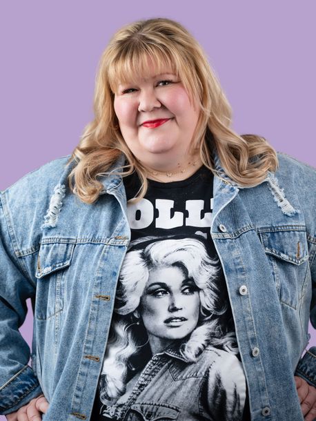 Woman in a denim jacket over a graphic tee, standing against a lavender background.