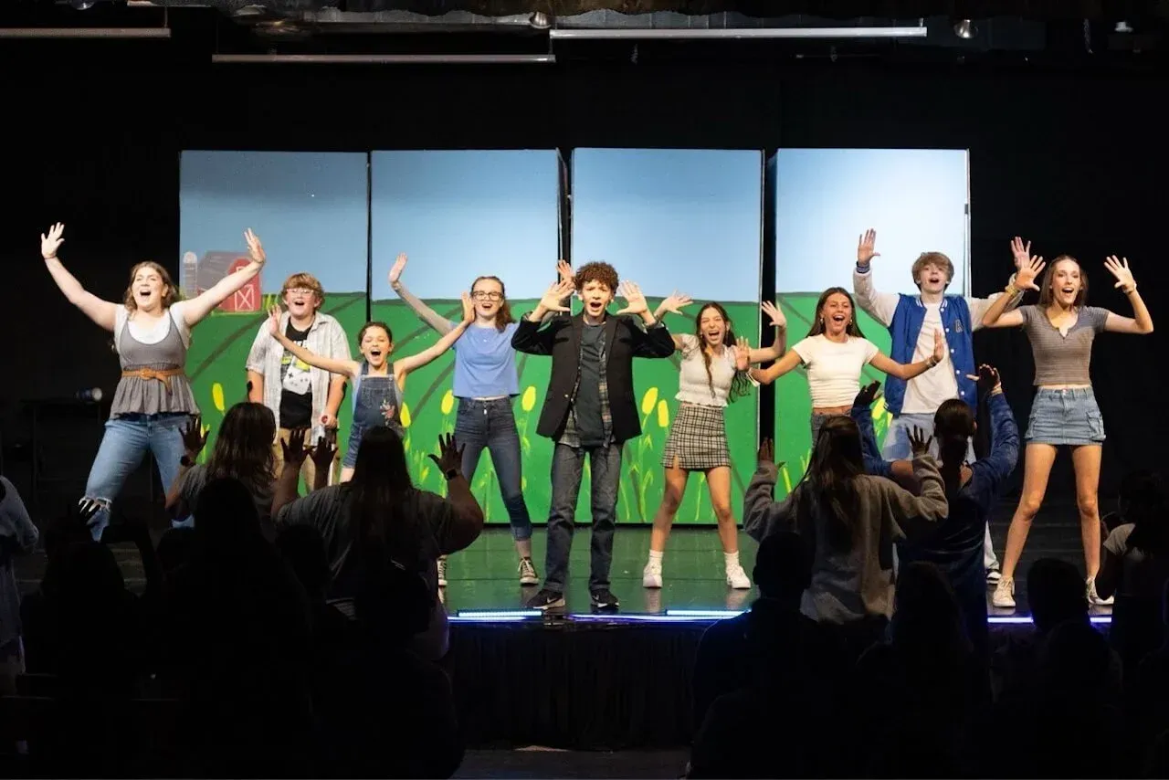 Group of performers on a stage, smiling and posing with arms raised under bright stage lights.