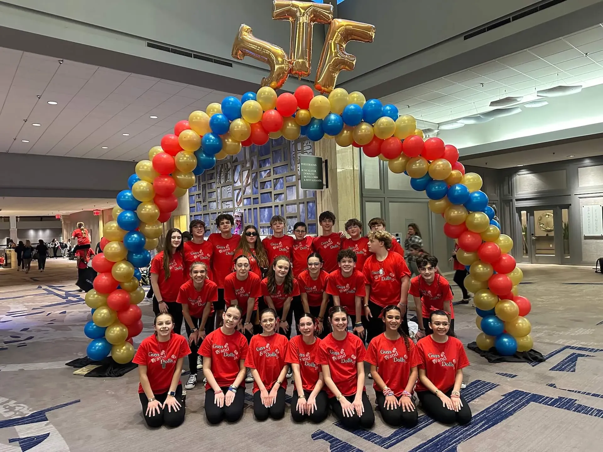 Group in red shirts posing under a red, yellow, and blue balloon arch in a hotel lobby