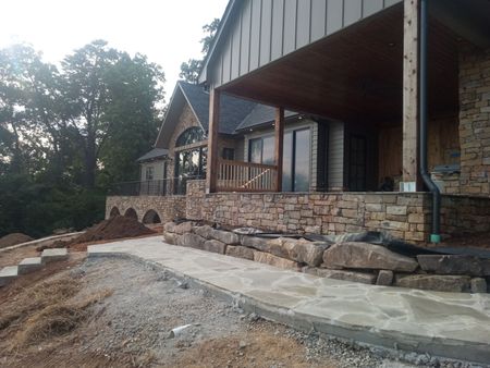 Stone house exterior with a covered porch and sloped walkway under construction, surrounded by trees