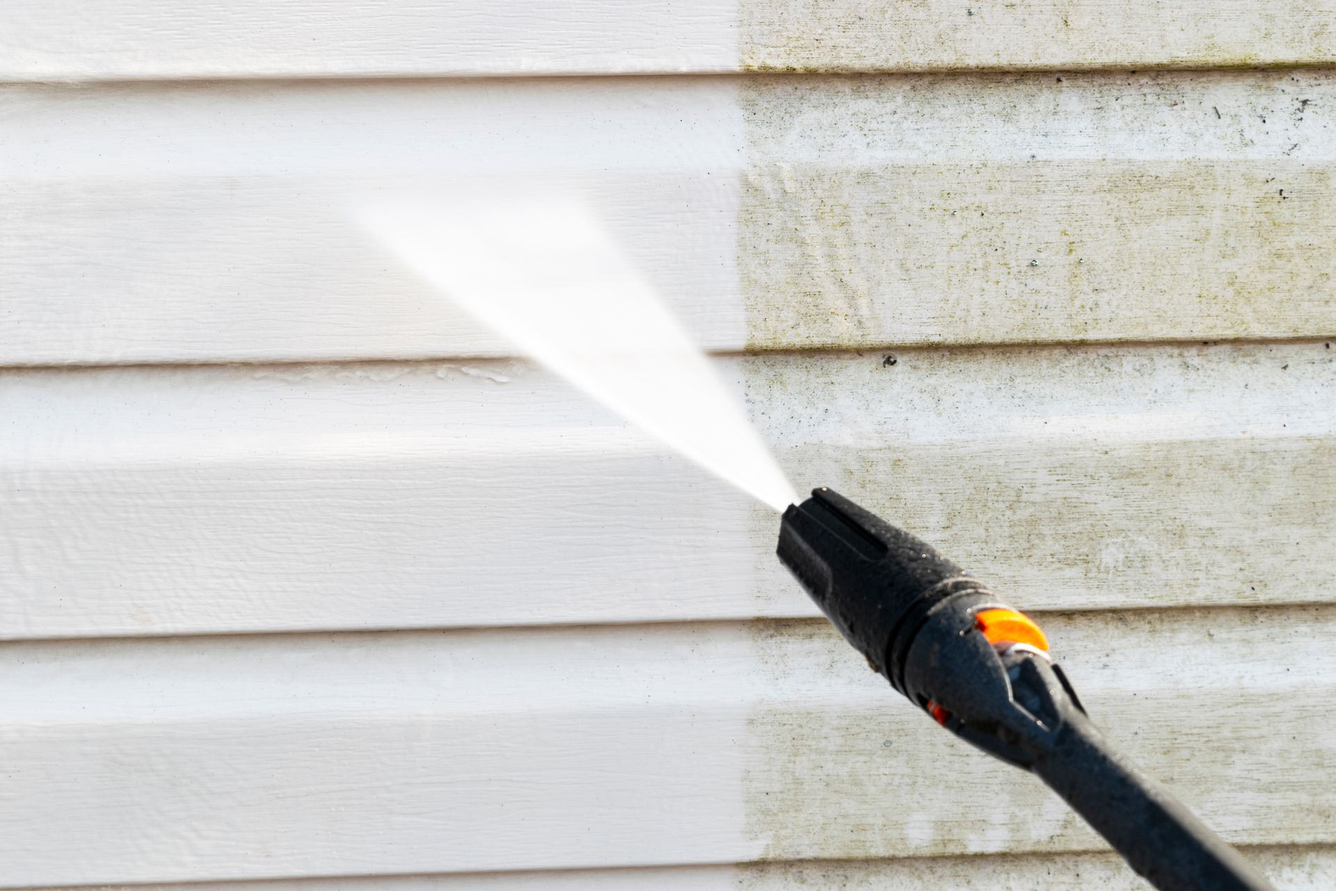 Pressure washing white vinyl siding beside a dirty wall with a spray nozzle