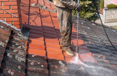 Worker pressure-washing a red tiled roof with water spraying over the shingles