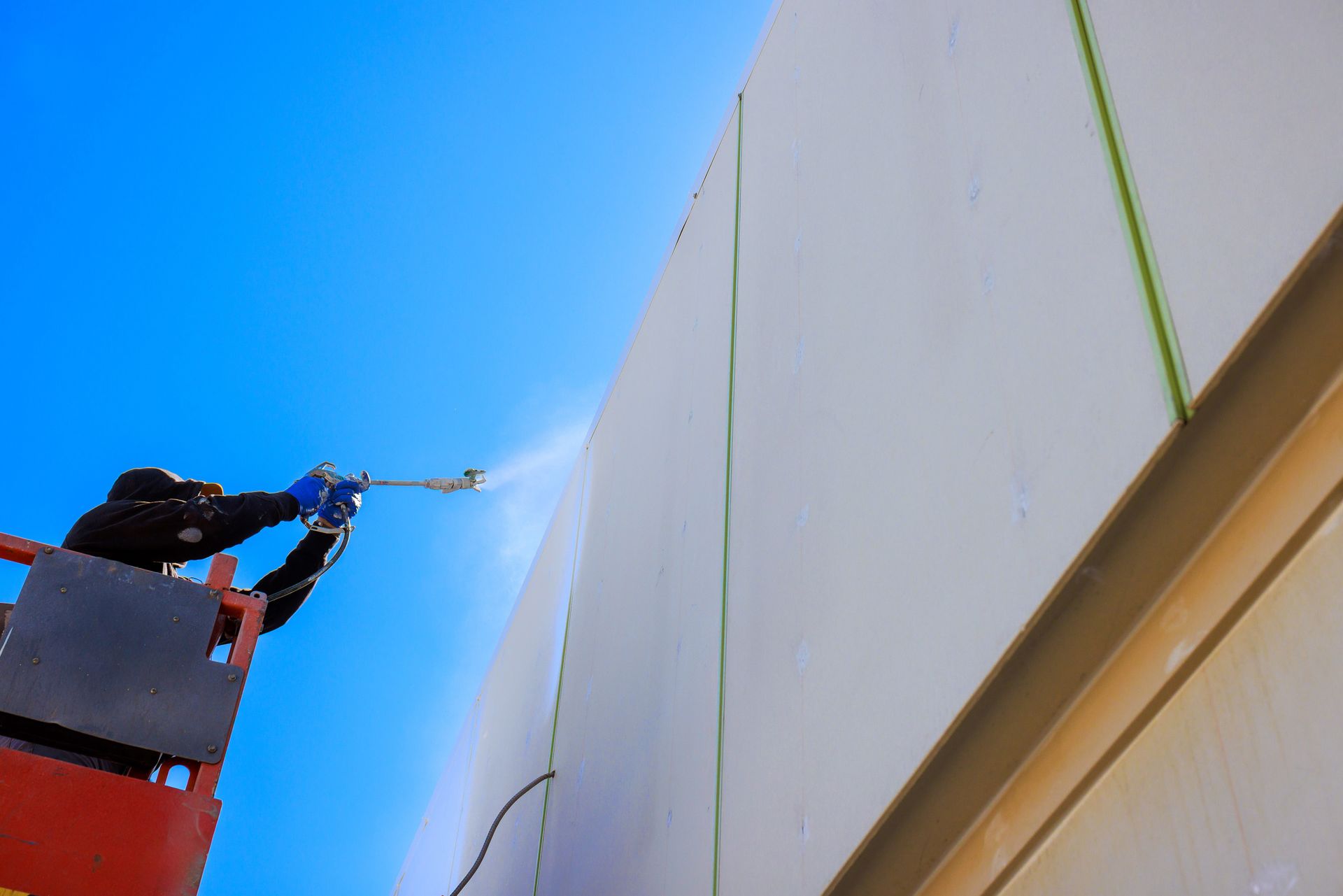 Worker pressure-washing a tall white building from a lift against a blue sky