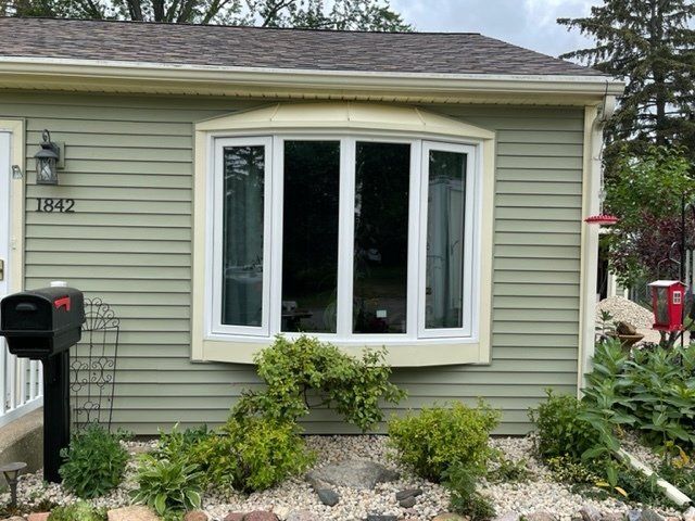 Front of a beige house with a bay window, black mailbox, and small shrubs in the garden.