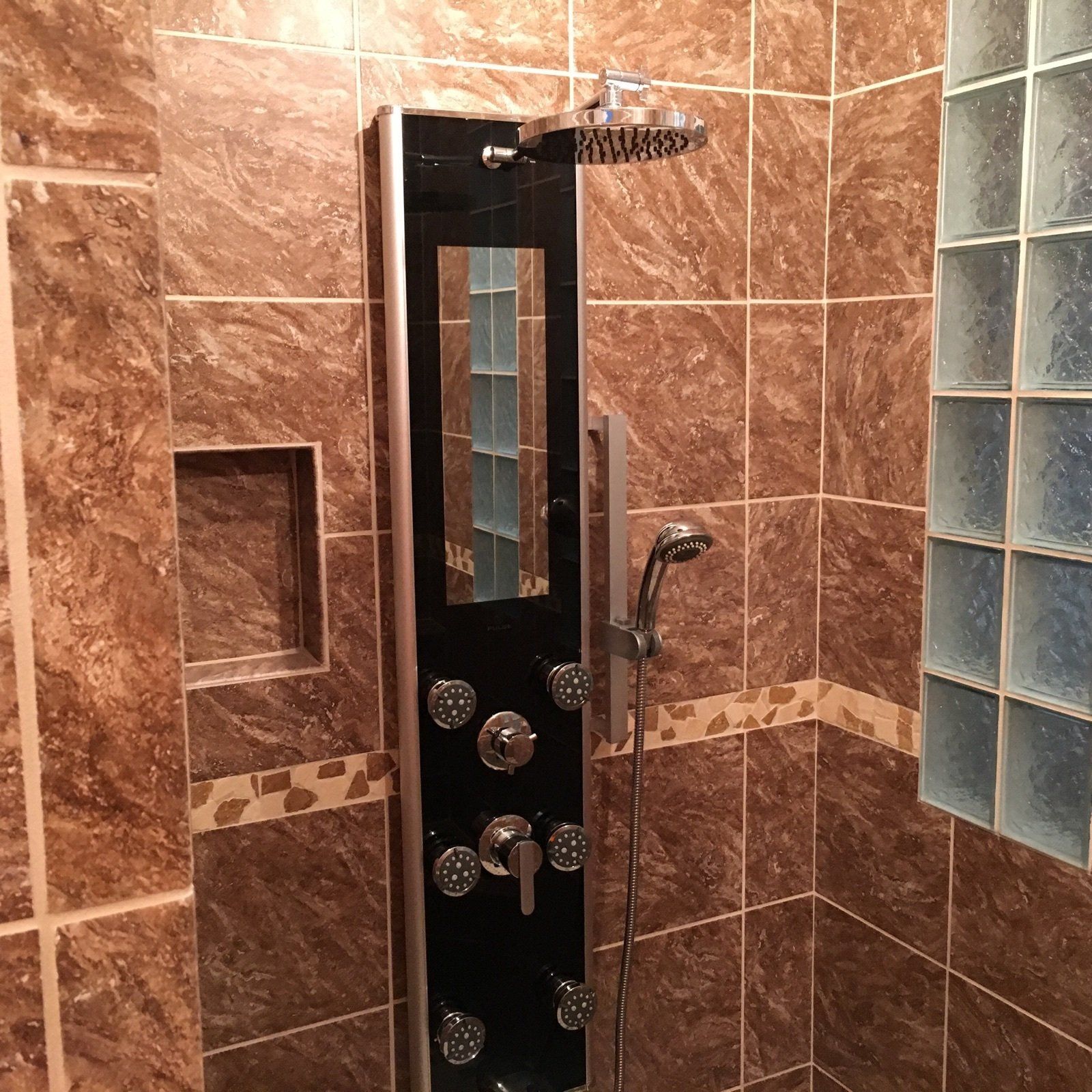 Tiled shower with a black wall-mounted control panel, handheld showerhead, and glass block window