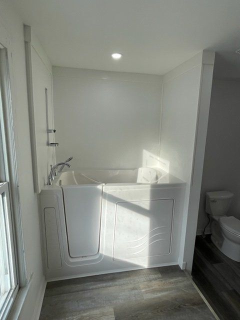 Bright white bathroom with a walk-in tub, gray wood floor, and toilet visible on the right.