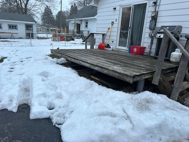 Snow-covered backyard deck beside a white house, with a ladder, red bucket, and footprints in the snow