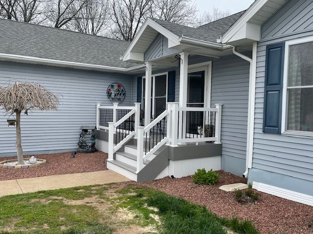 Front porch with white railings and steps on a gray house, featuring a small landscaped yard.