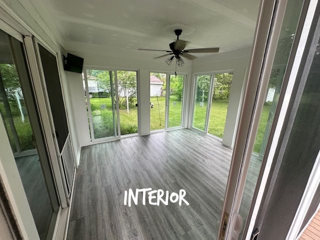 Empty sunroom with large glass windows, gray wood-look floor, ceiling fan, and green yard outside.