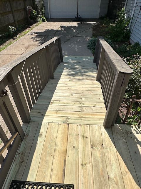 Wooden accessibility ramp with railings leading from a deck to a driveway, bordered by plants