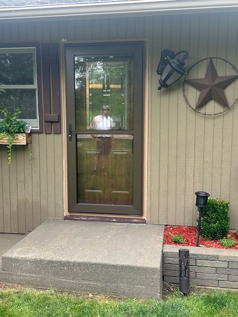 Front door entrance with concrete step, tan siding, hanging plant, star wall decor, and small flower bed