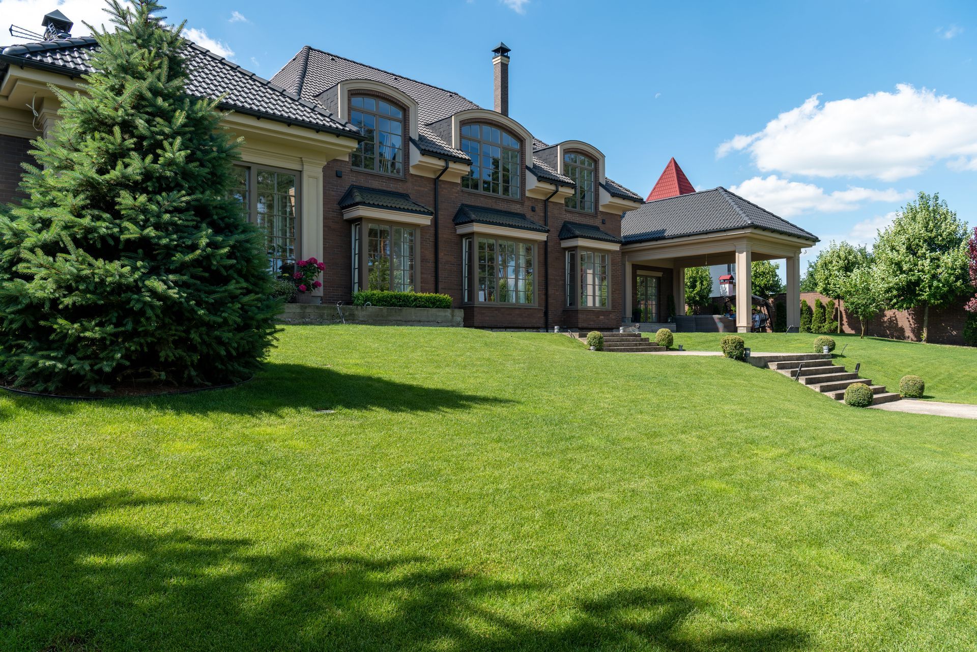 Large brick house with a manicured lawn, trees, and a covered patio under a bright blue sky