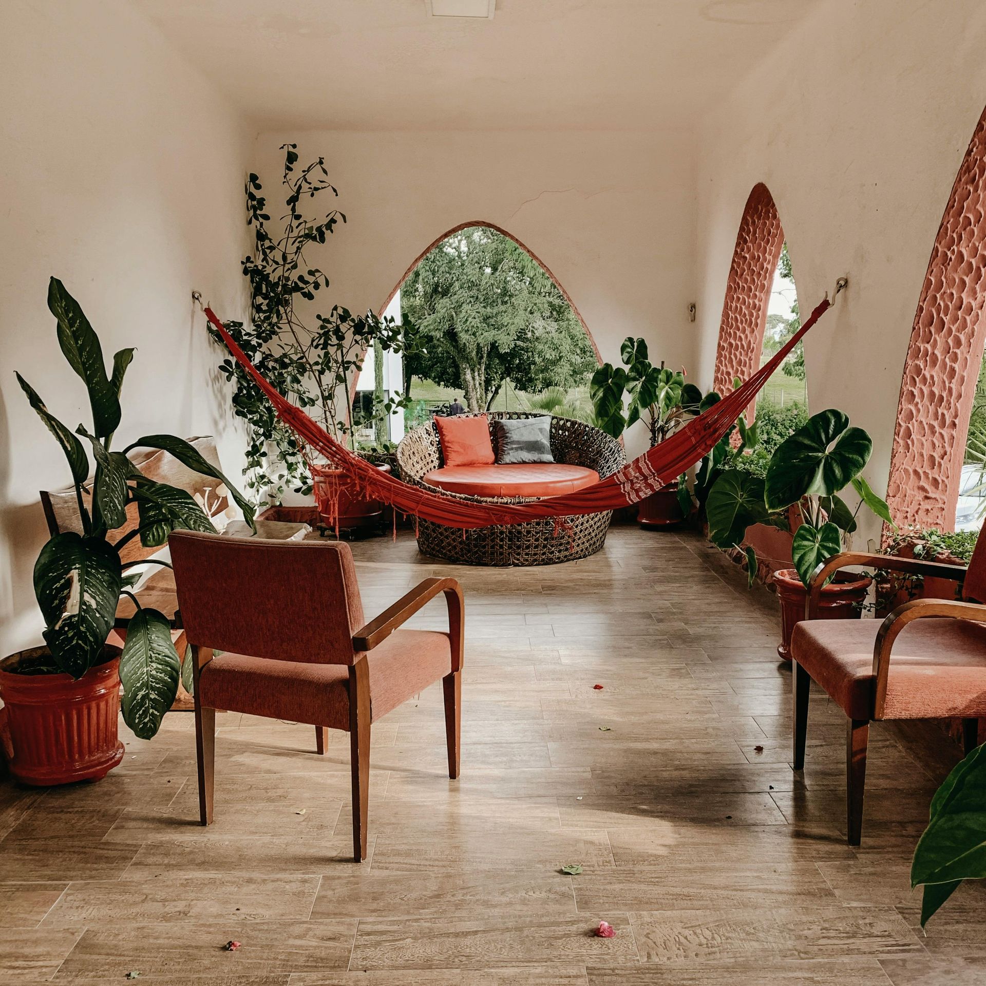 Covered porch with red hammock, chairs, and potted plants, opening to trees.