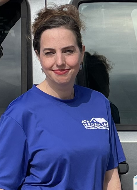 Woman wearing a blue shirt with a logo, smiling, standing outside near a vehicle.