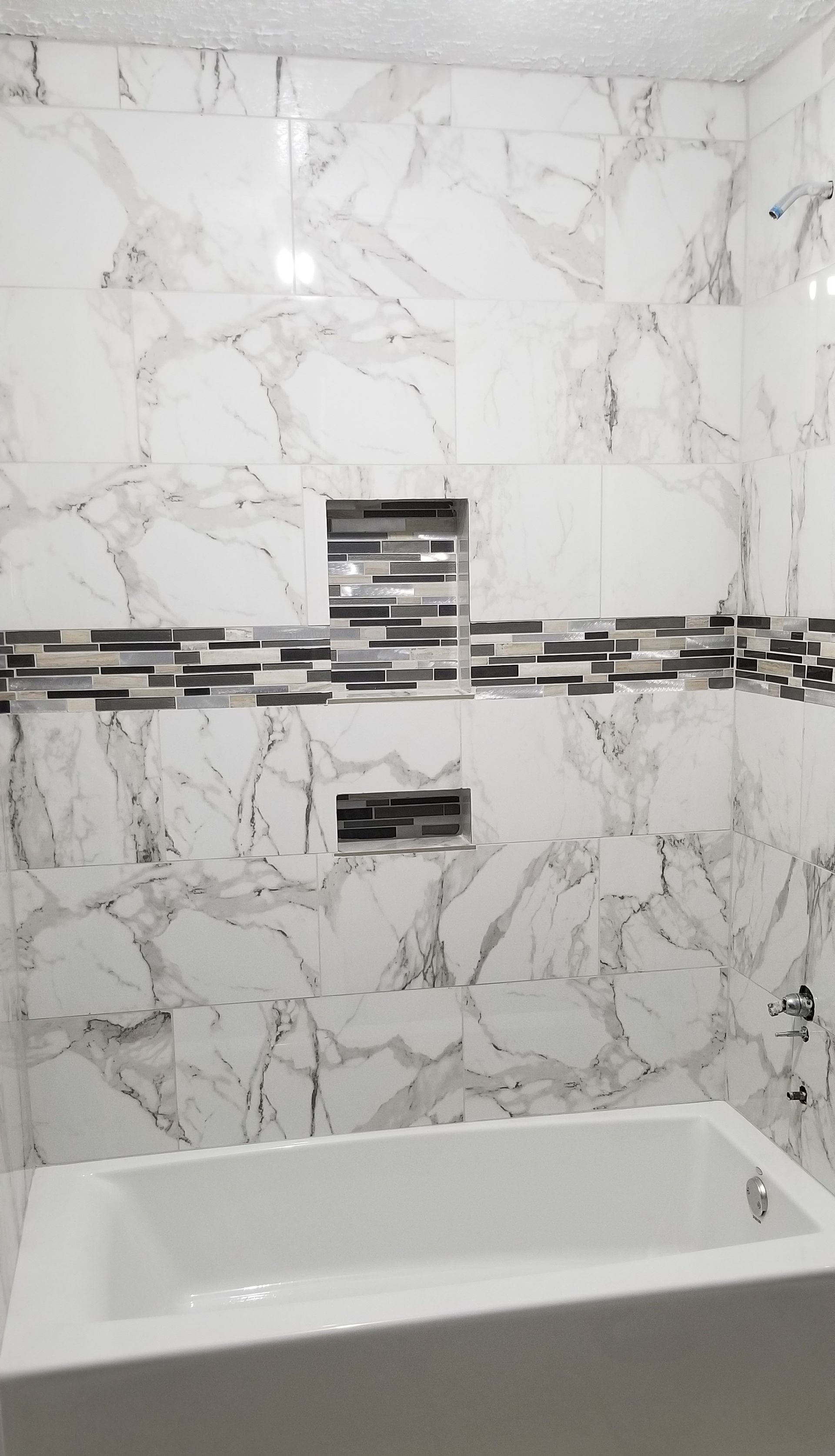 White marble-tiled bathroom with a bathtub. Decorative tile stripe and recessed shelves.