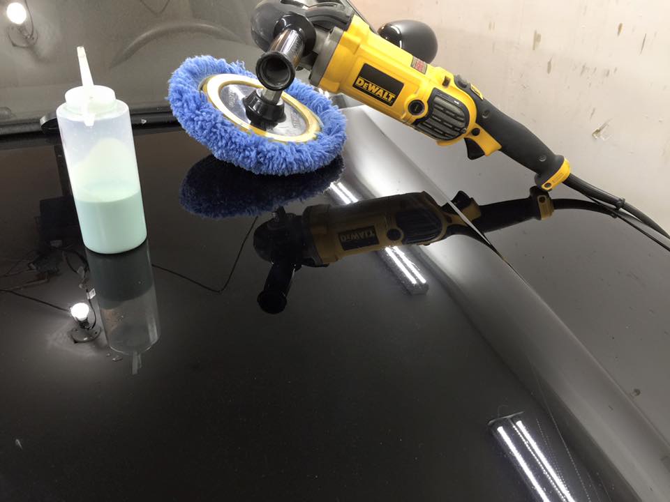 Yellow polisher with blue pad and liquid bottle on a glossy black car surface indoors