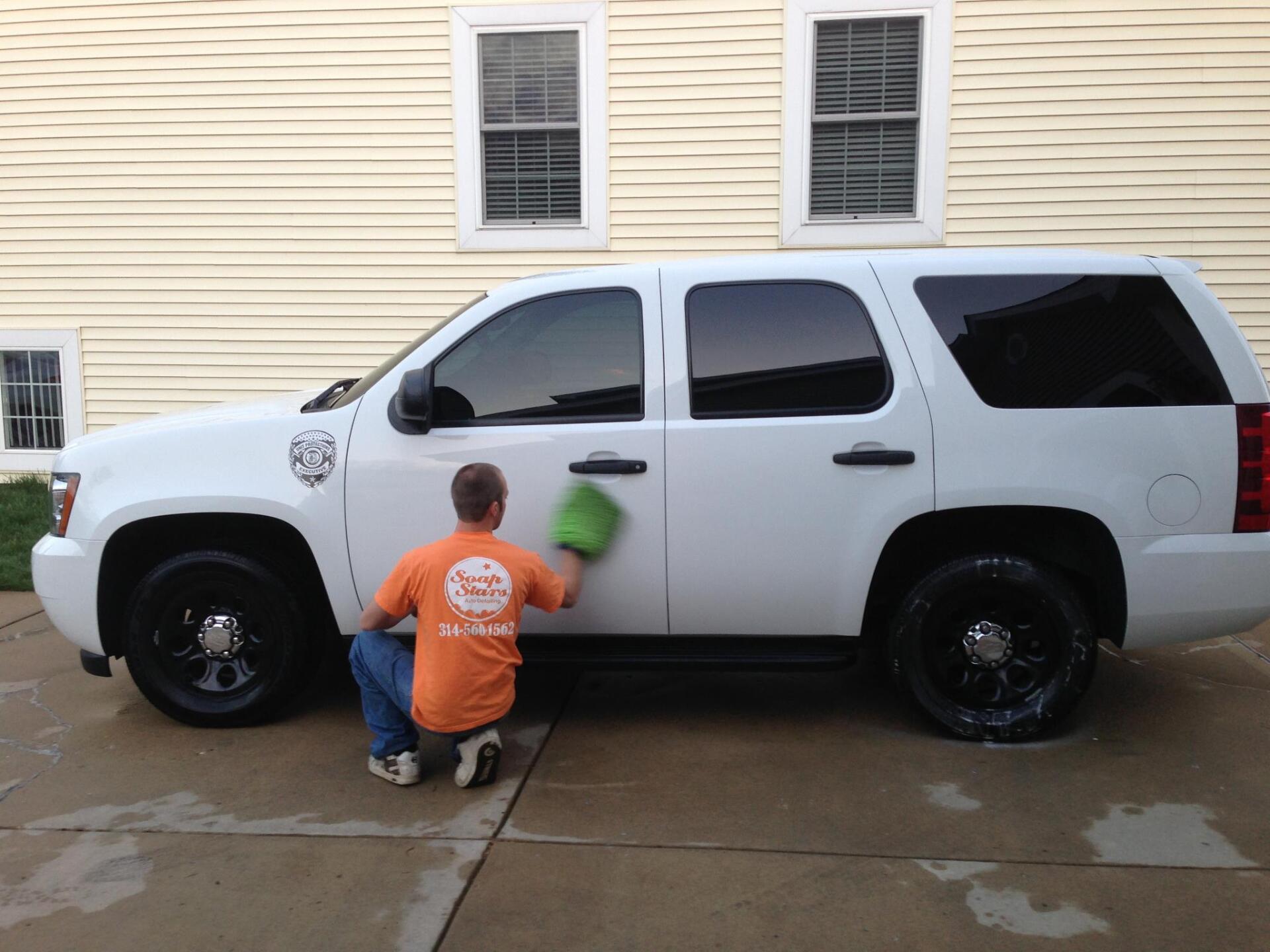 Person kneels by a white SUV, cleaning the driver door with a green sponge on a wet driveway.