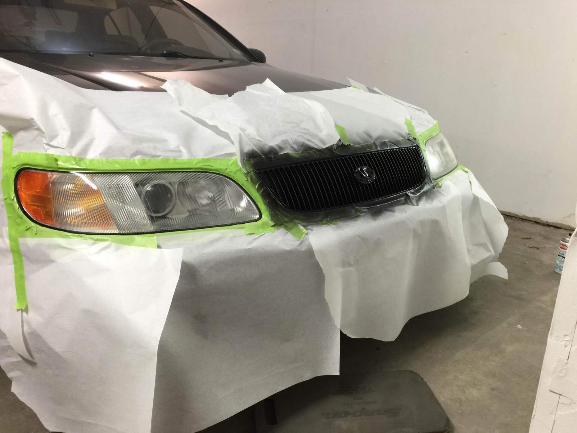 Car covered with masking paper and green tape in a garage, front bumper being painted or repaired