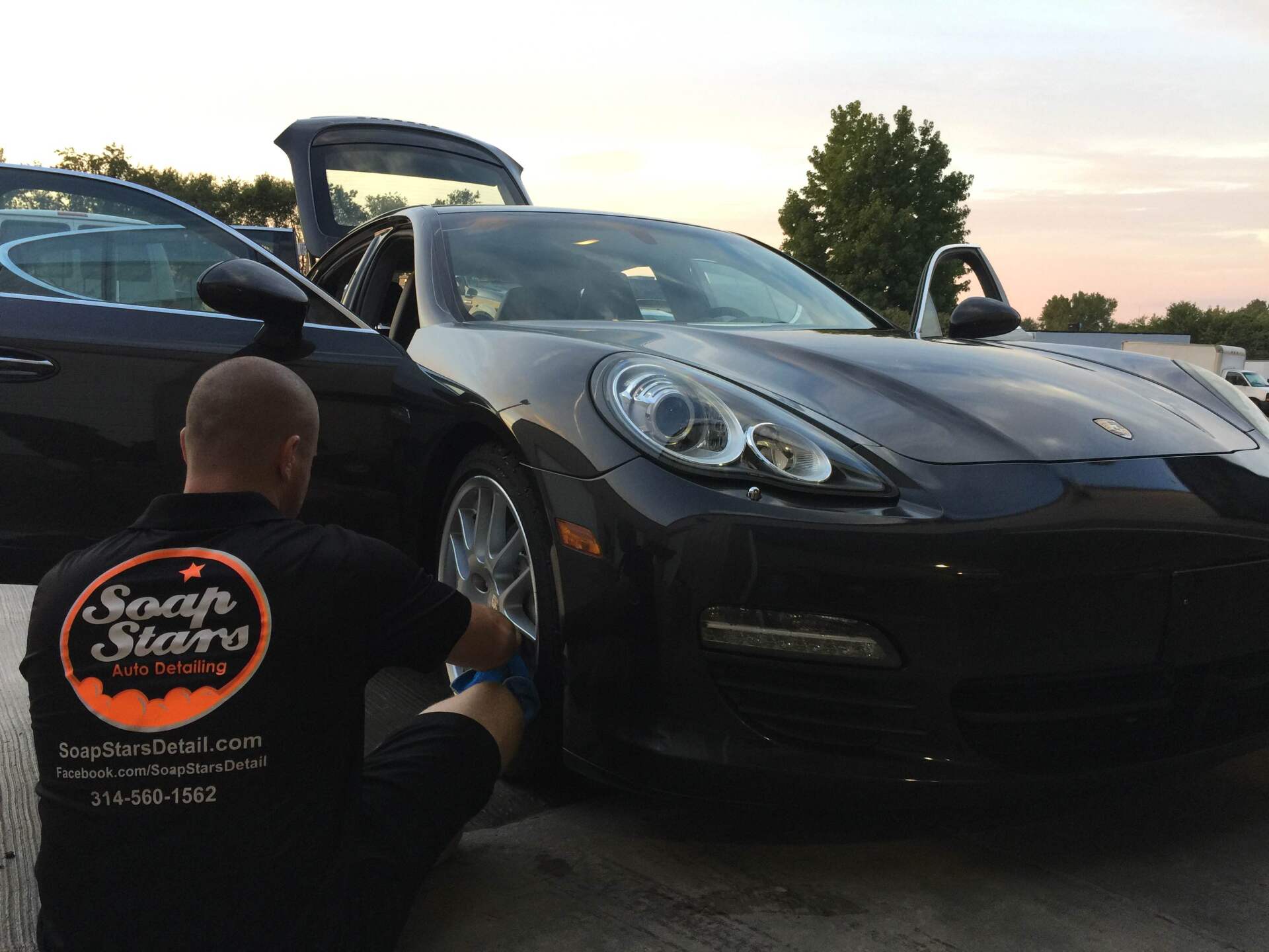 Person kneels by a black sports car with open doors at sunset, likely inspecting or washing it
