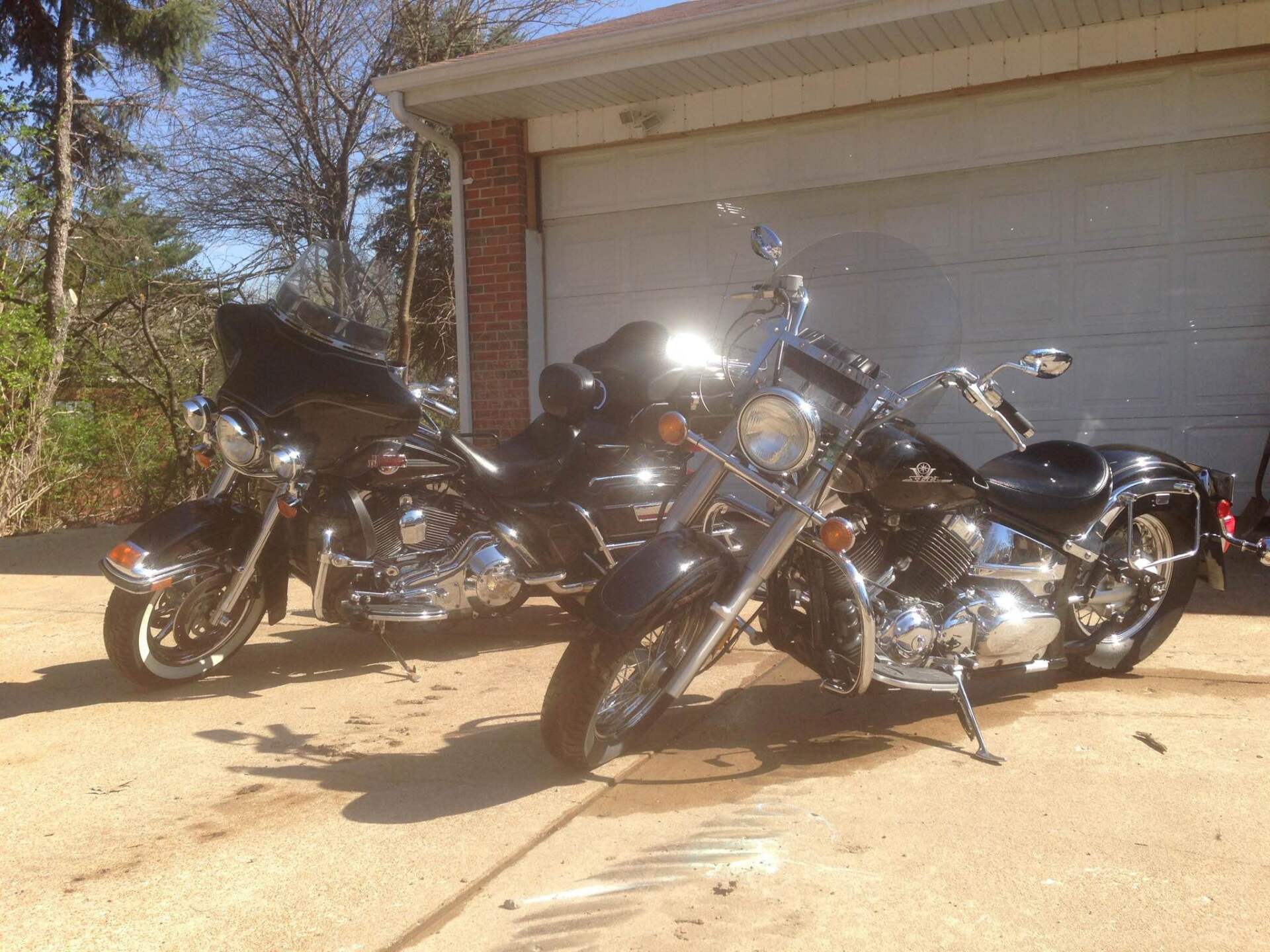 Two black motorcycles parked on a driveway in front of a garage and brick house.
