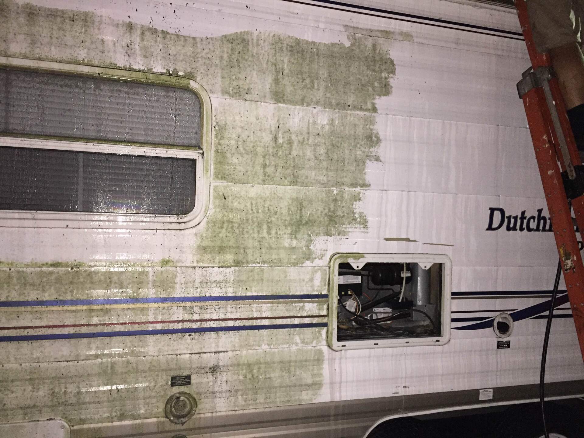 Dirty white RV side with green mildew stains, small window, and an open service hatch exposing wiring.