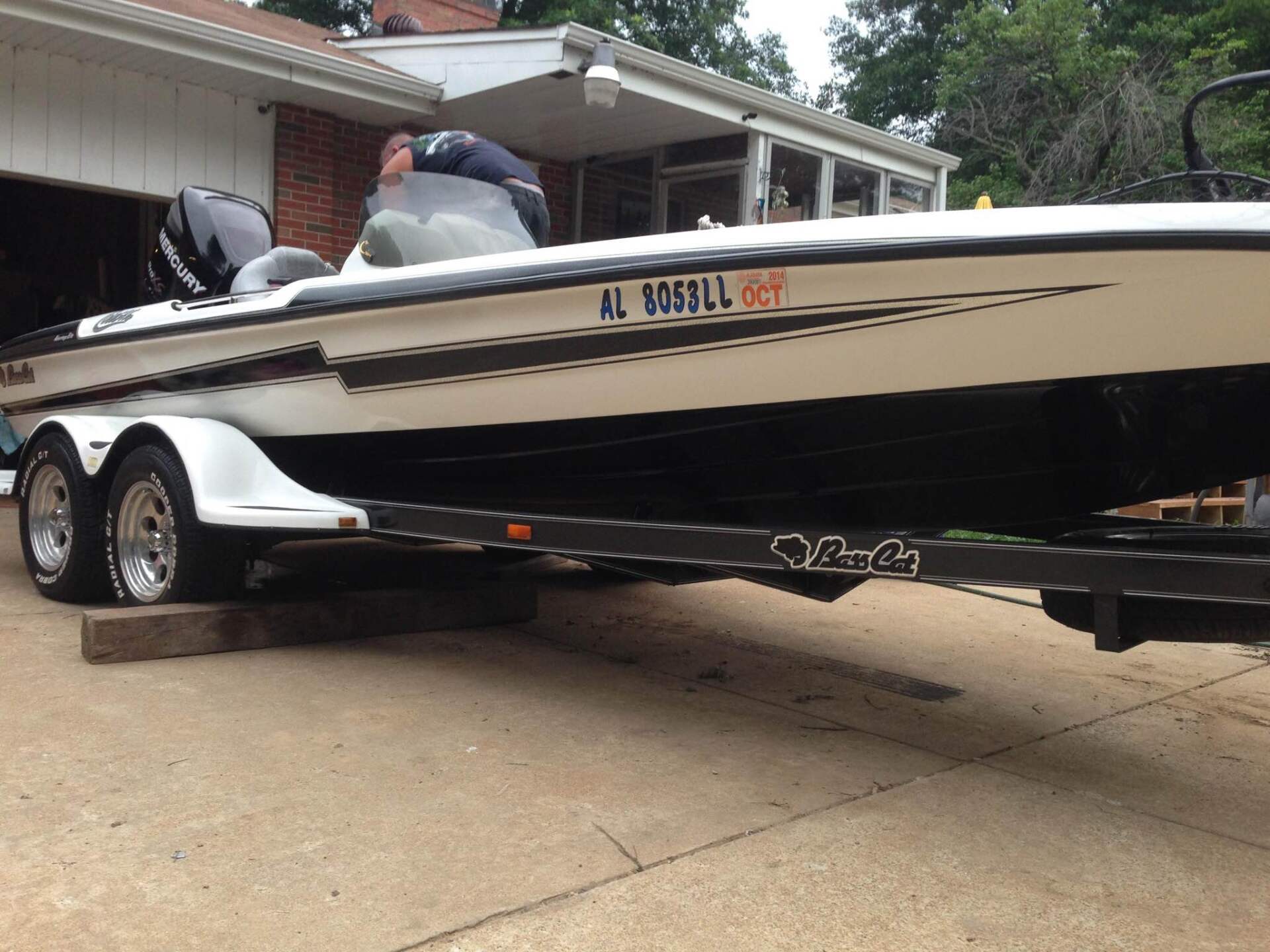 White and black speedboat on a trailer in a driveway, with a person working on it near a garage.