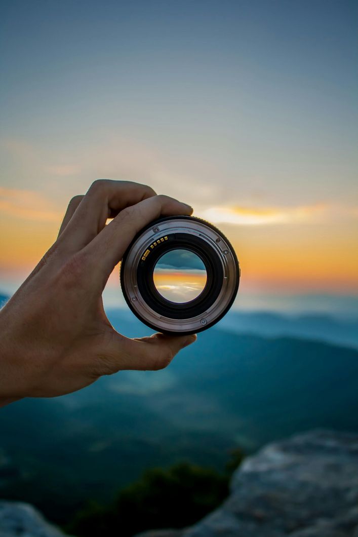 Hand holding a circular lens at sunset, framing a blurred mountain landscape and colorful sky
