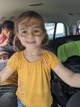 A little girl with her face painted is sitting in the back seat of a car.