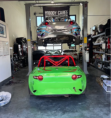 Green and red roll-bar Miata with a car on a lift in a garage; another car in the background.