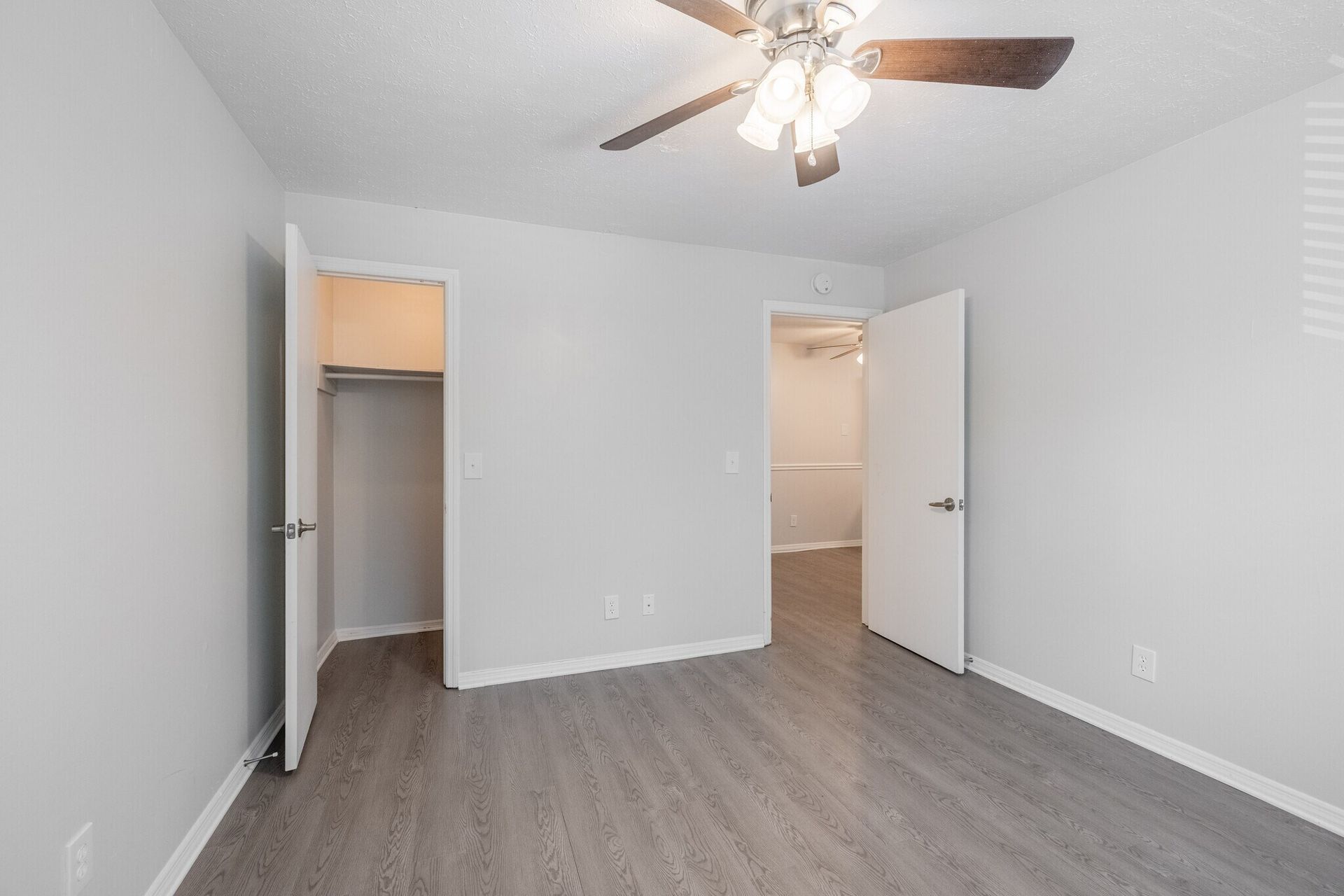 Empty bedroom with gray walls, wood-look flooring, ceiling fan, open closet, and two open doors at Glastonbury Woods Apartments.
