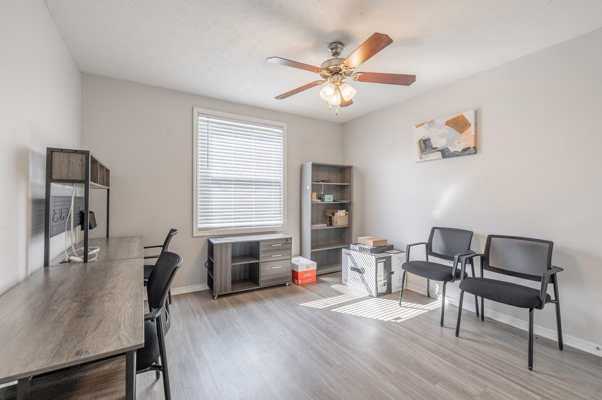 Office room with desk, chairs, shelving, window, ceiling fan, and art. Gray walls, wood-look floor at Glastonbury Woods Apartments.