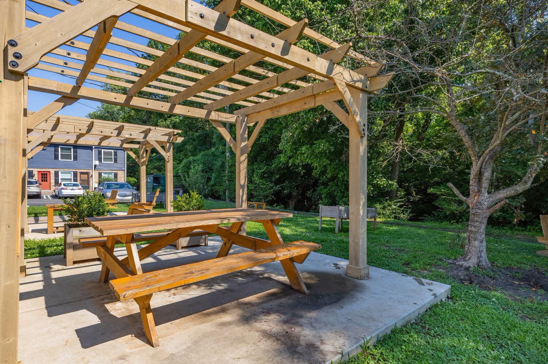 Wooden picnic table under a pergola on a concrete patio, with green trees and buildings in the background at Glastonbury Woods Apartments.