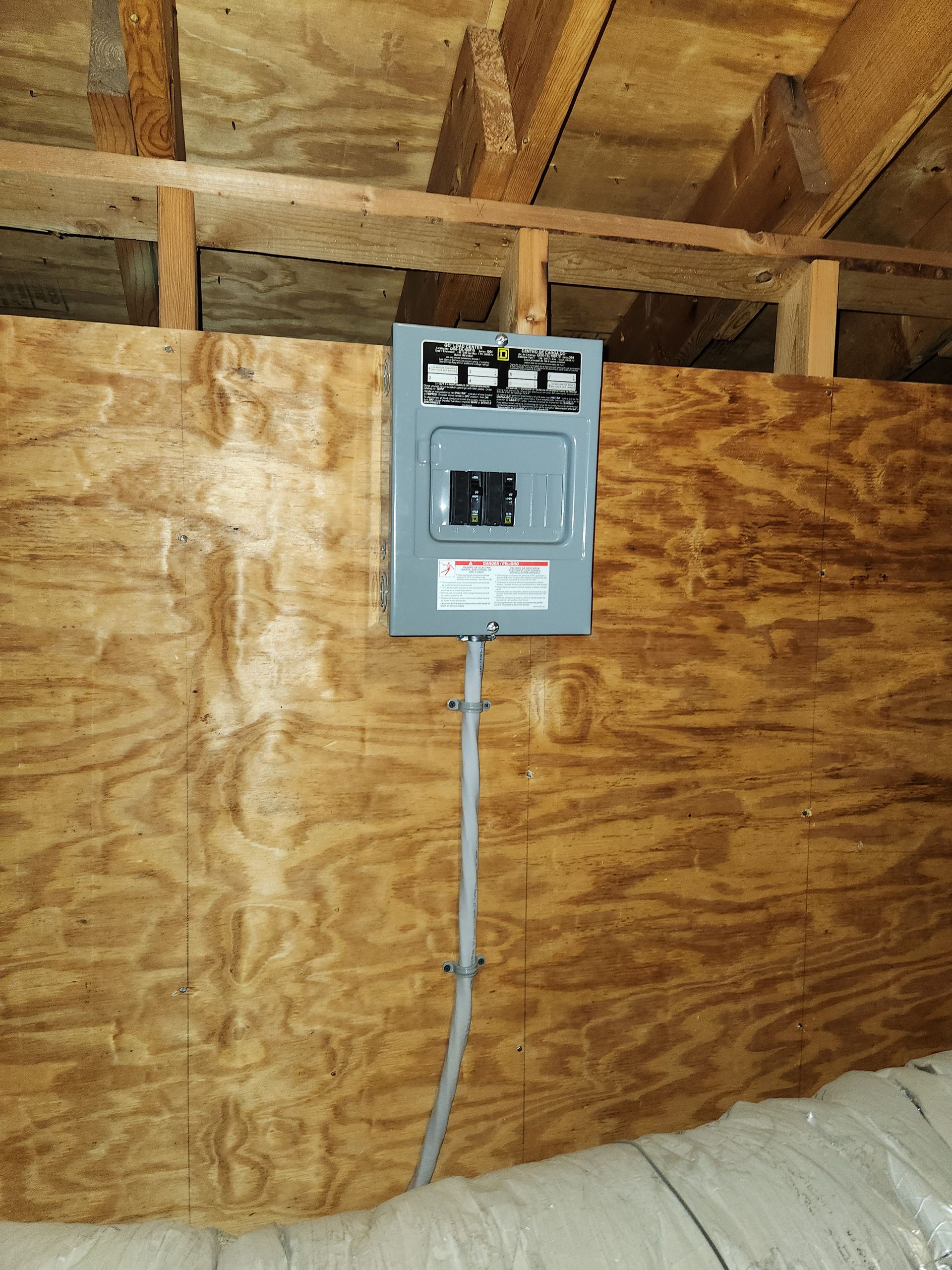Gray electrical breaker box mounted on plywood wall in an unfinished attic, with conduit running down.