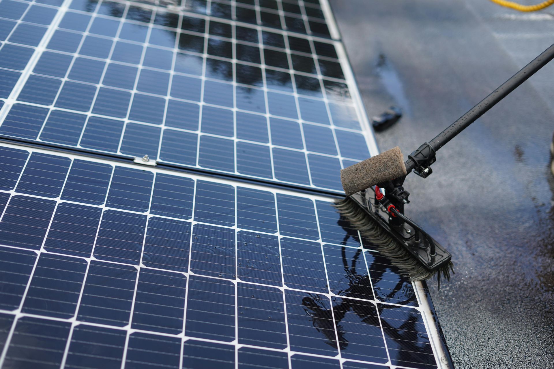 Solar panels being cleaned with a long-handled brush, reflecting a cloudy sky.