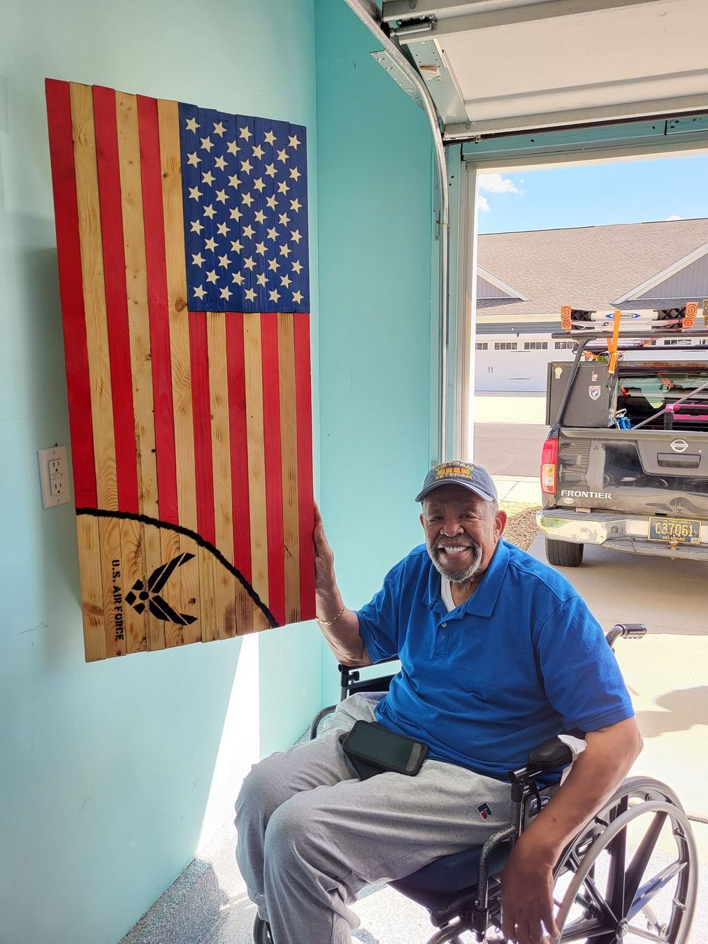 Man in wheelchair beside wooden American flag in a garage. He’s smiling.